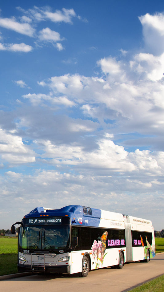 A long MTD hydrogen fuel cell bus with butterfly and flower graphics under a bright blue sky.